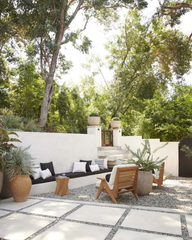 A bright, landscaped outdoor courtyard or patio with large pale square pavers set in gravel, a built-in white rendered bench with black cushions and pillows, wooden lounge chairs, and large ceramic planters. Short steps lead to a white rendered boundary wall and timber gate, surrounded by mature leafy trees and drought-tolerant planting.