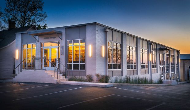 Twilight exterior view of a low-rise modern commercial building, likely a renovated or purpose-built community or office space. The long facade combines light painted brick, extensive floor-to-ceiling window bays with dark metal frames, a projecting entrance canopy, exterior wall lights, and concrete steps with metal handrails. Warm interior lighting contrasts with the cool blue evening exterior. A small adjoining older structure is visible at the left, with paved parking in the foreground.