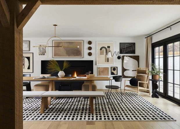 A bright, refined open-plan living and dining area framed by substantial exposed timber posts and beams. The room features pale timber flooring, a long timber dining table with bench seating, a large black-and-white patterned rug, a dark media console, framed abstract artwork, sculptural lighting, a lounge chair, indoor plants, and expansive black-framed glazed doors. A small fireplace glow adds warmth to the calm, curated interior.