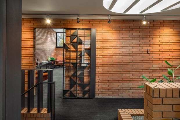 Interior view of a contemporary industrial-style space featuring exposed red brick walls, dark carpeted flooring, black metal-framed glazing and a black metal geometric stair or display structure. A curved slatted ceiling feature with integrated lighting spans the upper right, while brick-built planters and sparse greenery appear in the foreground.