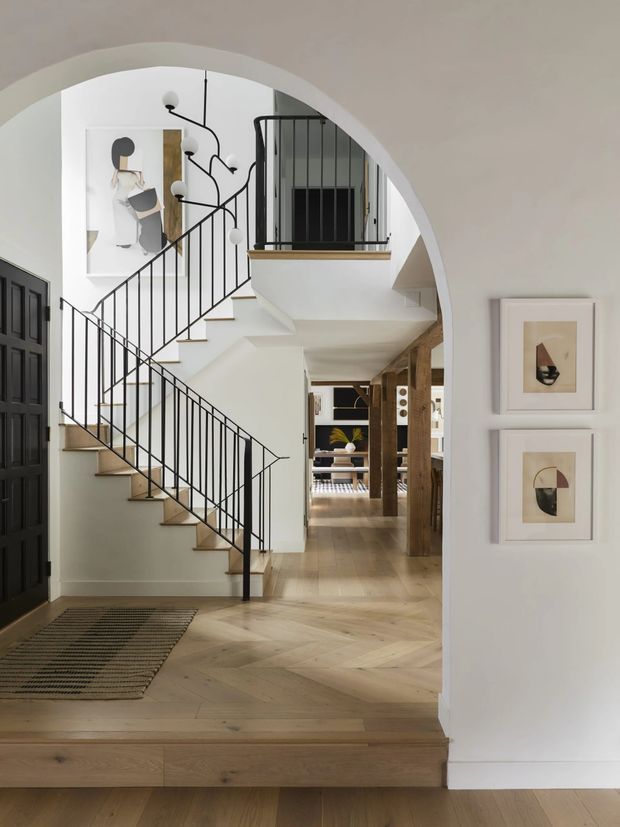 Bright, refined residential entry hall viewed through a large curved archway, featuring a sculptural split staircase with slim black metal balustrades, pale timber herringbone flooring, white walls, framed abstract artwork, and a glimpse into a living area beyond. A dark paneled door and timber structural details add contrast.