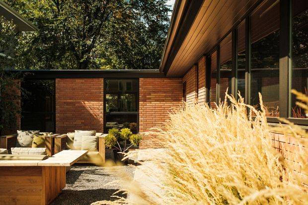 A warm, low-slung brick house exterior surrounding a landscaped courtyard or patio. The facade combines horizontal reddish brickwork, dark-framed glazing, and a timber-lined soffit beneath a shallow pitched roof. An outdoor seating area with a built-in timber element and light cushions sits beside gravel landscaping and ornamental grasses, creating a sheltered, private residential courtyard.