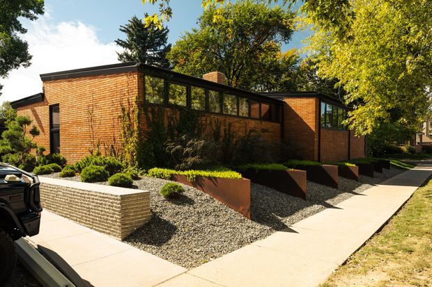 Exterior view of a low, single-storey brick building with a dark flat roof, continuous high-level windows, and a landscaped frontage. The building is set along a concrete pedestrian path, with gravel garden beds, rust-coloured steel planters, clipped shrubs, and mature trees. The architecture has a strong mid-century institutional or modernist character, with horizontal proportions and restrained detailing.