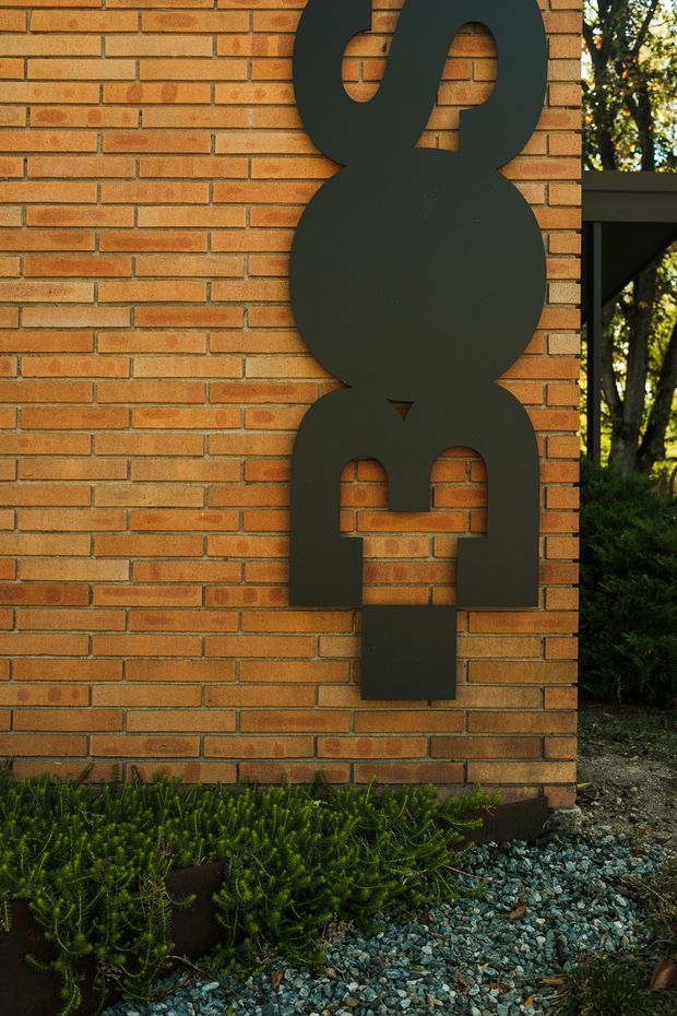 Close-up exterior view of a warm orange brick wall featuring a large, vertically stacked matte-black abstract metal sculpture or signage element. Low planting and blue-grey gravel sit at the base, with a dark roof or canopy structure and greenery visible at the right edge.