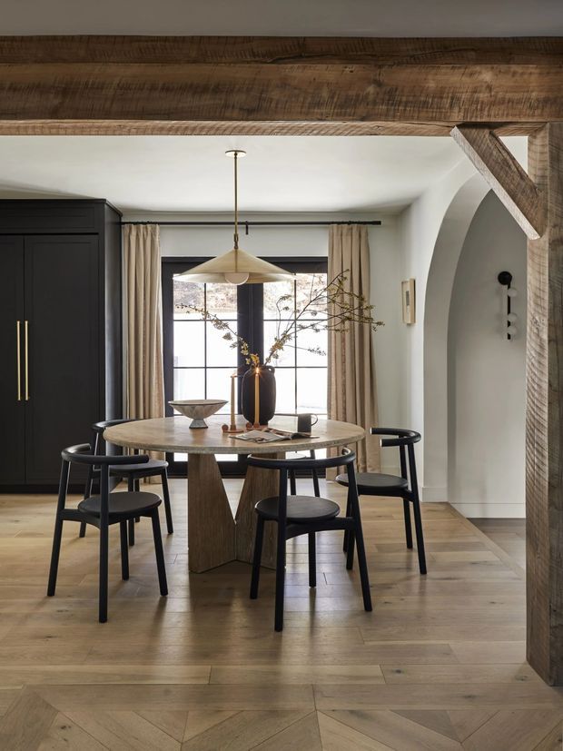 A refined dining room with a round sculptural timber dining table and four black dining chairs, positioned in front of a large gridded window with beige curtains. The room features warm timber flooring, dark built-in cabinetry, a brass-toned pendant light, white walls, and prominent exposed timber beams framing a curved arched opening.