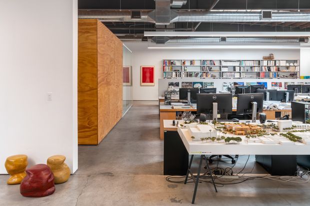 An open-plan commercial design studio or office with exposed services overhead, polished concrete flooring, timber-clad partition volumes, extensive shelving filled with books and samples, workstations with computer monitors, and a large architectural model table in the foreground. Bright white walls and colourful sculptural seating add visual accents.