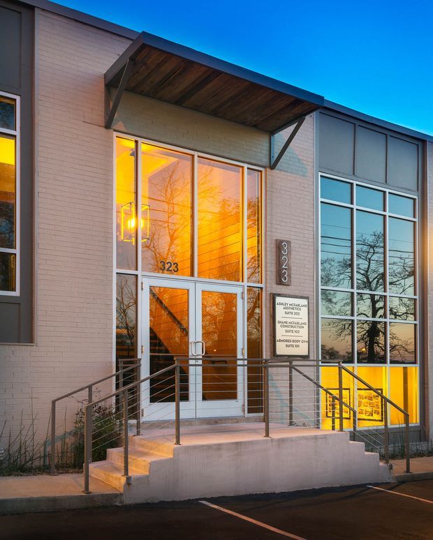 Exterior view of a modern commercial building entrance at dusk, featuring a glazed double-door entry, large metal-framed windows, pale brick walls, a projecting timber-lined canopy, and stainless-steel handrails. Address numbers and a professional office sign are visible beside the entrance.