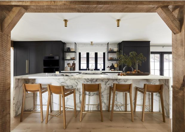 A sophisticated kitchen viewed through a substantial timber-framed opening. The room features a large marble-look island with a waterfall edge and seating for five, matte black floor-to-ceiling cabinetry, open shelving, brass hardware and fittings, timber bar stools, light timber flooring, and black-framed windows and doors. The palette is warm and neutral with strong black-and-white contrast, complemented by natural greenery and styling.