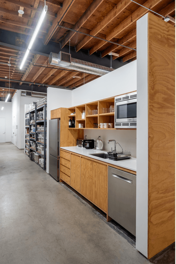 A compact commercial kitchenette or break room set within an open industrial workspace. The space features warm plywood cabinetry and shelving, white walls, a white benchtop, integrated stainless-steel dishwasher, sink, microwave, refrigerator, and small appliances. Exposed timber ceiling joists, visible ductwork, suspended linear lighting, concrete flooring, and adjacent metal storage shelving create an industrial workshop or warehouse character.