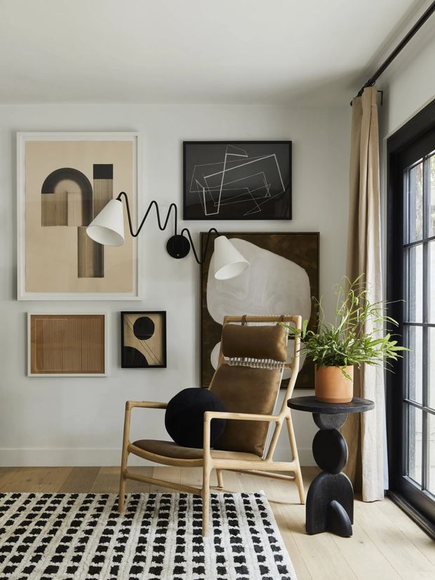 A stylish, light-filled living room corner with a warm neutral palette. A tan upholstered lounge chair and matching wooden-framed armchair sit beside a sculptural black side table topped with a potted plant. Framed abstract artwork, a wall-mounted black-and-white lamp, full-height cream curtains, a black-framed glazed door or window, pale timber flooring, and a bold black-and-white patterned rug complete the interior.