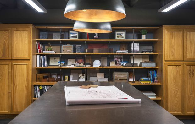 A sophisticated home office or studio workspace with a large dark worktable in the foreground, architectural drawings spread across it, extensive open shelving filled with books and design objects, and tall timber storage cabinetry on both sides. The room has a dark ceiling, exposed industrial-style lighting, and two large metallic pendant lights with warm interiors.