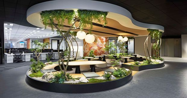 A contemporary commercial interior featuring an open-plan office arranged around large organic, oval-shaped planter islands. The space has integrated seating, indoor trees and lush planting, sculptural cream ceiling forms with suspended globe lights, dark ceiling panels, grey flooring, workstations, and a colourful feature artwork wall. The design creates a biophilic, refined workplace environment.
