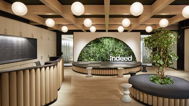 A sophisticated contemporary commercial interior, likely a reception or lounge area, featuring a curved upholstered reception counter or bar at left, extensive cream cabinetry, a dark textured feature wall, and a large circular living-wall feature with the “indeed” logo. The space has a warm timber coffered ceiling with abundant spherical pendant lights, light timber flooring, curved dark seating, sculptural stools, and indoor trees integrated into rounded furniture.