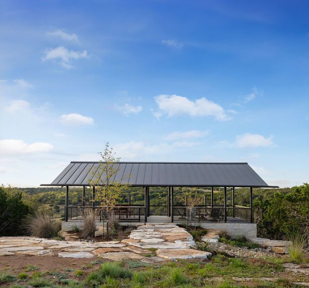 Single-storey pavilion-like building set in a rugged landscaped site, with a long dark standing-seam pitched roof, extensive black-framed glazing, and an open terrace or living area visible behind the glass. A natural stone path and rock landscaping lead toward the structure, with low scrubland surrounding it under a bright blue sky.