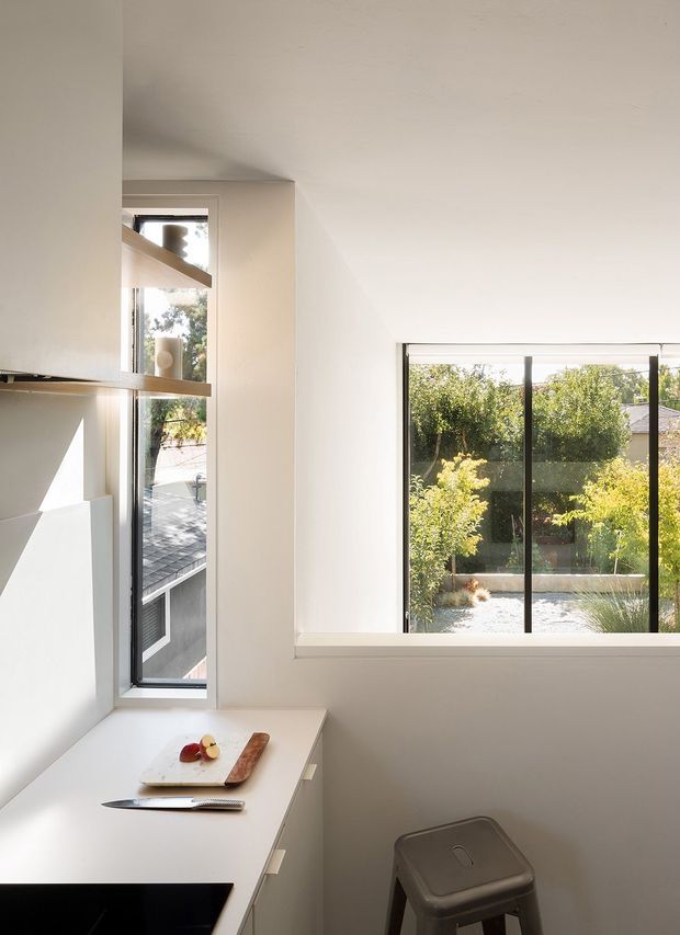 Bright, minimalist kitchen corner with white walls and white benchtops, a narrow vertical window and a wide black-framed horizontal window overlooking a landscaped outdoor area. A cutting board, fruit, knife and a simple metal stool are visible, with strong natural sunlight creating geometric shadows.
