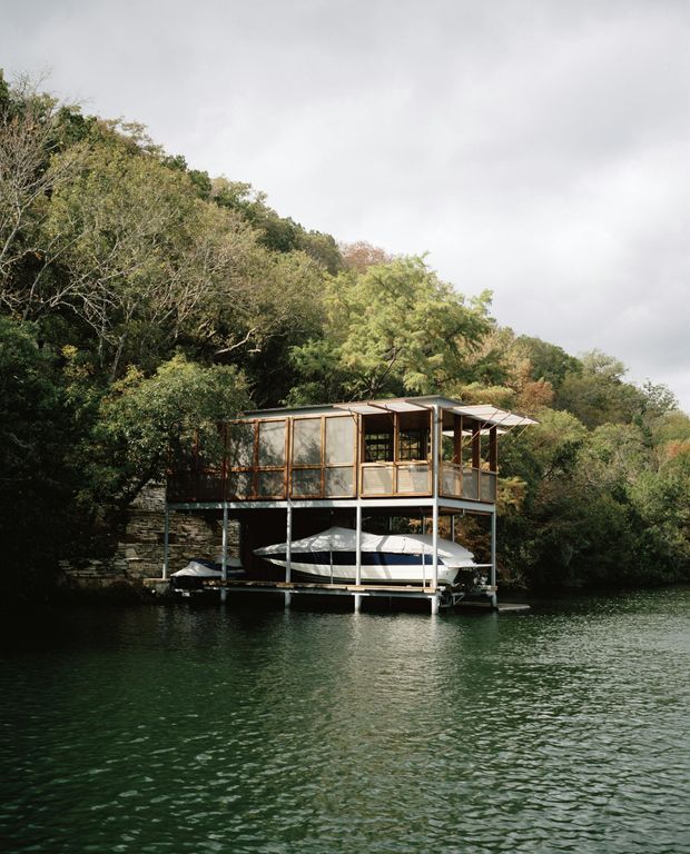 A compact elevated waterside structure stands on a floating or stilted platform above a calm, tree-lined body of water. The upper level has timber-framed screened glazing, open-sided areas, a small covered deck, and a lightweight pitched roof. A white boat is stored beneath the structure, with dense woodland and rocky shoreline surrounding the scene.