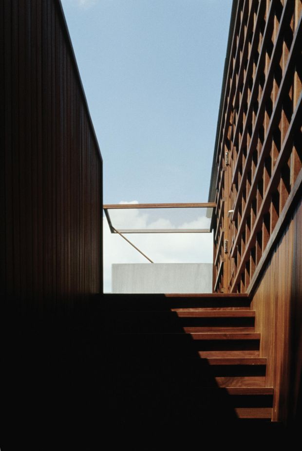A dramatic architectural view looking up a narrow exterior stairway or stepped passage between two tall building volumes. Dark timber-clad walls frame the left and right sides, while the right facade features a repeating open geometric screen or structural grid. Horizontal beams span the gap overhead beneath an open sky, creating strong linear shadows and a sculptural, modern composition.
