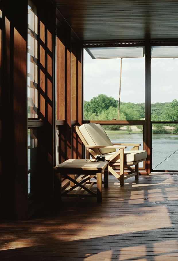A sunlit enclosed outdoor room or glazed balcony overlooking a river or waterfront landscape. The space features extensive dark timber wall panelling, timber decking, large framed glazing, a slatted ceiling, and simple timber lounge chairs with cream cushions. Strong diagonal shadows create a warm, tranquil atmosphere.