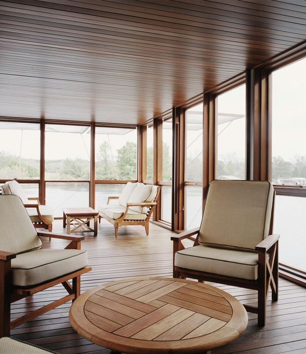 A bright enclosed sunroom or lakeside sitting area with expansive floor-to-ceiling glazing, warm timber-framed windows, and a timber-lined ceiling and floor. Several mid-century-inspired upholstered lounge chairs surround a round timber coffee table, with a calm natural landscape and water visible outside.