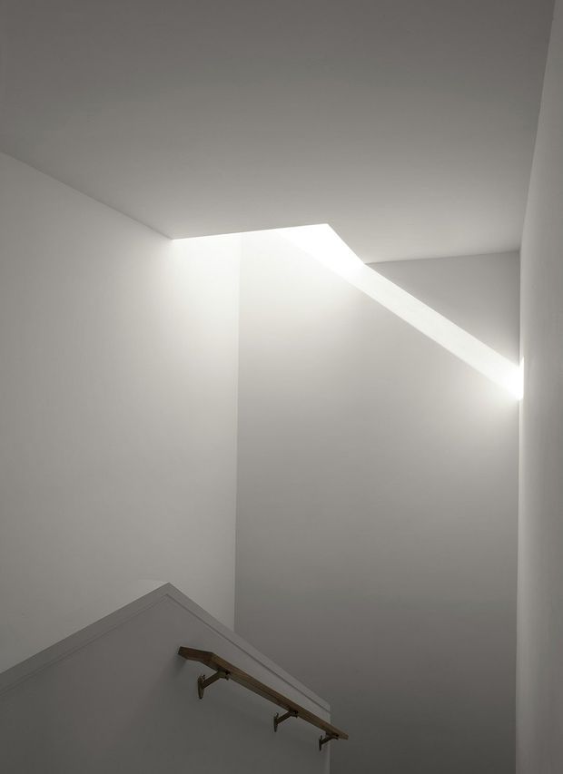 Minimal, monochromatic interior view looking upward in a stairwell or stair landing. Smooth white plaster walls and ceiling are intersected by a dramatic angular skylight or roof opening that floods the space with daylight. The lower portion shows a simple stair handrail with dark metal brackets and a timber-toned rail. The composition is architectural and highly pared back, emphasizing geometry, light, and shadow.