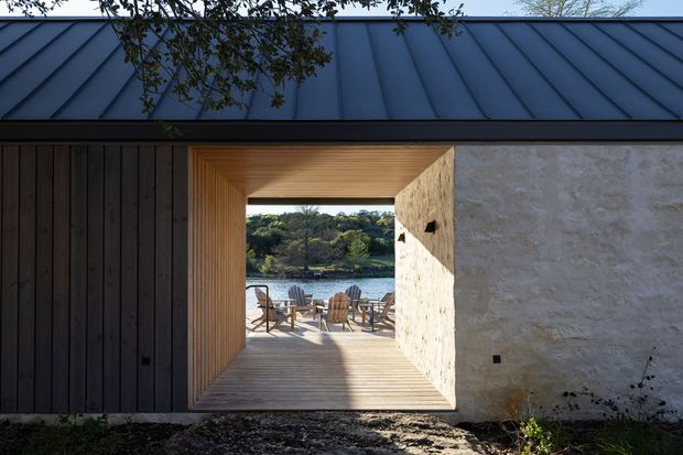Exterior architectural view of a contemporary pavilion or house with a dark standing-seam metal roof, vertical dark timber cladding, and a pale textured masonry wall. A timber-lined rectangular passage frames a deck with outdoor seating and a water view beyond.