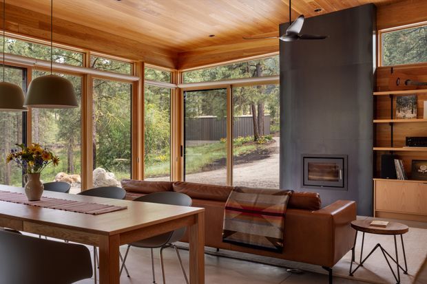 A warm, wood-lined open-plan living and dining area surrounded by expansive windows overlooking a wooded setting. The room features a timber dining table, pendant lighting, a leather sofa with a patterned throw, a dark fireplace wall with a built-in fireplace, and open timber shelving and cabinetry.