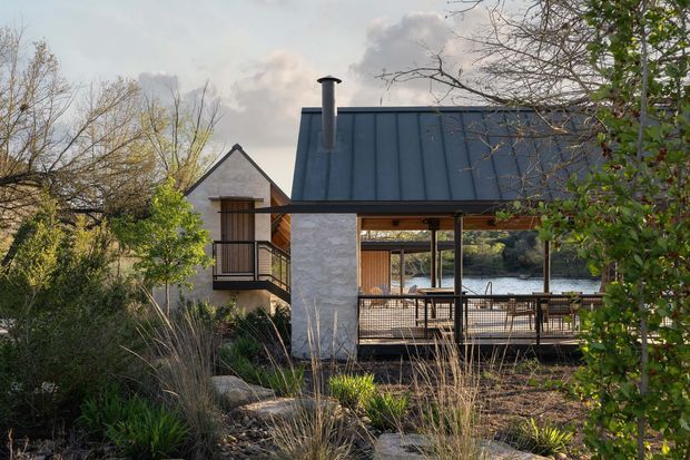 Exterior view of a contemporary residential pavilion beside a body of water, surrounded by established landscaping. The building features a steep dark standing-seam metal roof, pale masonry walls, timber detailing, expansive open glazing, and a covered outdoor dining or entertaining deck with railing. A smaller gabled volume connects to the main pavilion.