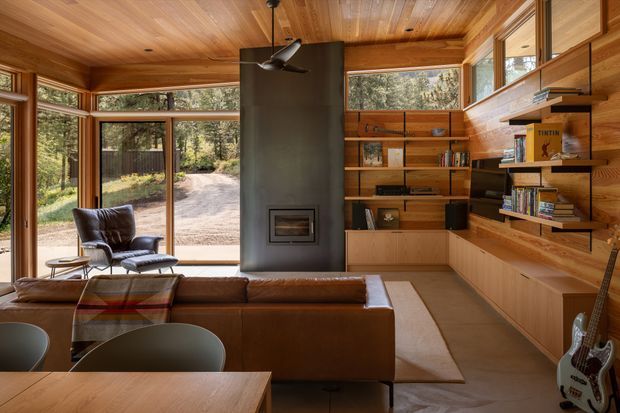 Warm, timber-lined open-plan living room with floor-to-ceiling glazing overlooking a wooded driveway. A freestanding dark fireplace forms a central partition, with built-in timber shelving and cabinetry along the right wall, a leather sectional sofa, lounge chair, dining table, and guitar. The space has a strong connection to the surrounding landscape and abundant natural light.