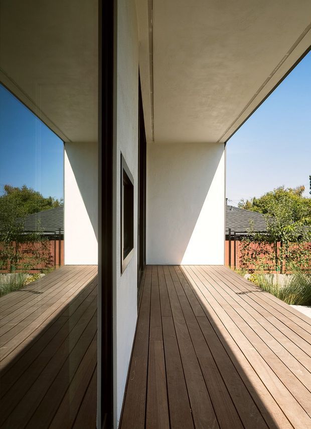 A contemporary covered timber deck or terrace viewed beside a large reflective glazed opening. The space has warm brown timber decking, white rendered walls and soffits, dark slim window or door frames, and strong geometric sunlight and shadow. A landscaped garden and neighbouring rooflines are visible beyond the terrace.