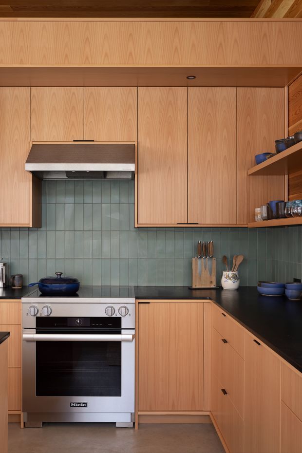 A compact, modern kitchen with extensive vertical-grain timber cabinetry, dark benchtops, and a muted blue-green tiled splashback. A freestanding stainless-steel range cooker sits beneath a concealed rangehood, with open timber shelving displaying cookware and utensils. The warm timber ceiling and cabinetry contrast with the cool backsplash and dark work surfaces.