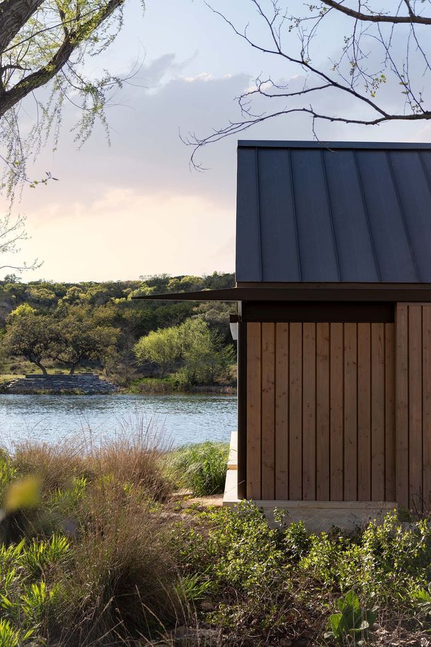 A close exterior view of a compact modern building beside a calm river or lake, surrounded by dense natural vegetation and mature trees. The structure features dark vertical metal wall cladding, warm vertical timber boarding, a simple projecting roof edge, and a low, rectilinear form. Soft late-day light creates a quiet connection between the building and its wooded waterfront setting.