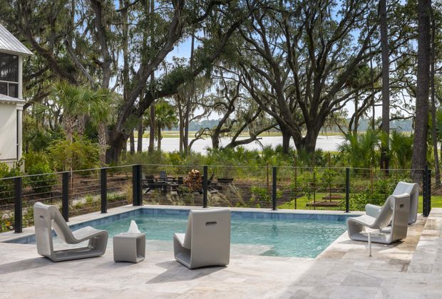 A contemporary outdoor pool terrace overlooking a landscaped waterfront setting. The compact rectangular pool is surrounded by pale stone paving and sculptural white outdoor lounge chairs, with a dark wire-and-post balustrade, tropical planting, mature trees, and water visible beyond. A portion of the adjacent house appears at the left edge.