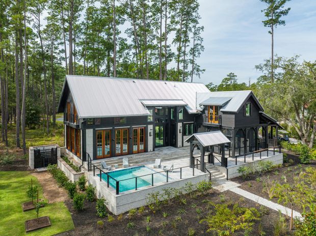 Exterior aerial view of a large contemporary timber-clad residence set within a wooded landscape. The home has multiple pitched gabled roof forms with light metal roofing, dark exterior cladding, extensive glazed doors and windows, elevated decks and covered outdoor areas. A terrace with a small turquoise swimming pool, railings, stairs, planted beds and newly landscaped grounds occupies the foreground.