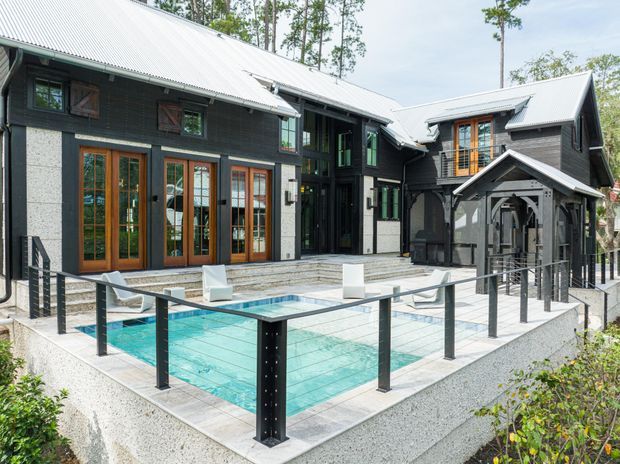 Exterior view of a substantial dark-clad, multi-wing house arranged around a raised tiled terrace and swimming pool. The home features steep corrugated metal gabled roofs, contrasting light masonry or rendered wall sections, extensive timber-framed glazed doors and windows, and a covered entry structure. Black metal pool balustrades enclose the foreground terrace.