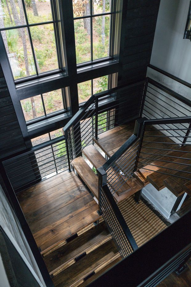 A dramatic multi-level stairway and landing viewed from above, featuring dark timber wall cladding, large black-framed windows, warm timber stair treads and landings, and black metal railings with horizontal cable infill. The glazing overlooks a wooded landscape, while a white structural wall provides contrast.