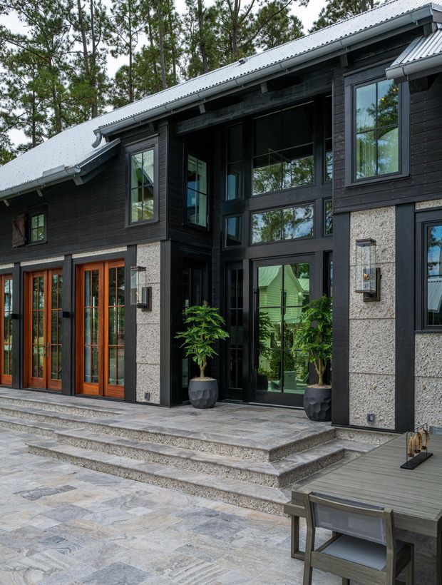 Exterior view of a contemporary dark-clad house set among trees, featuring a steeply pitched light metal roof, large black-framed glazing, timber-framed glazed entry doors, textured light stone wall panels, and a broad paved terrace with steps and outdoor dining furniture.