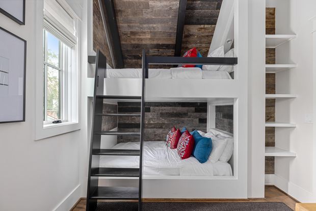 Compact bunk-room interior with two built-in sleeping levels framed in white joinery. A black metal ladder and guard rail provide access to the upper bunk, while both bunks have white bedding and colourful accent cushions. The walls and ceiling feature dark, weathered timber-look boards, contrasted with white painted walls, trim, and open shelving. A window brings in natural light, creating a practical, cozy sleeping space.