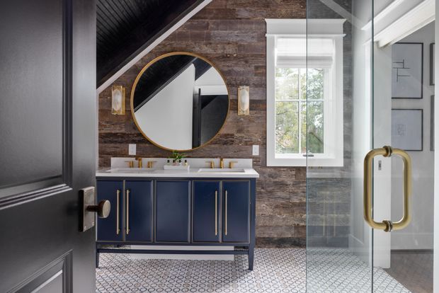 A refined bathroom vanity area viewed through a partially open dark door and glass shower screen. The room features a navy-blue double vanity with brass pulls, white countertop and basins, a large round brass-framed mirror, dark timber-look wall cladding, patterned geometric floor tiles, and a white-trimmed window. A brass shower handle and glass enclosure are prominent on the right.