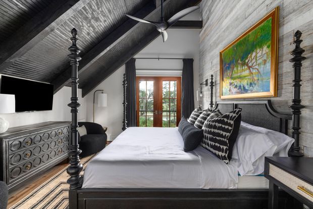A dramatic, highly styled bedroom with a pitched, exposed-beam ceiling and dark timber framing. The room features a large black four-poster bed dressed in white linen and patterned black-and-white cushions, a matching dark dresser, dark curtains, a ceiling fan, and a timber-framed glazed door at the end. Grey textured wall cladding and ceiling boards create a moody, rustic-industrial backdrop, complemented by a colourful framed landscape painting.