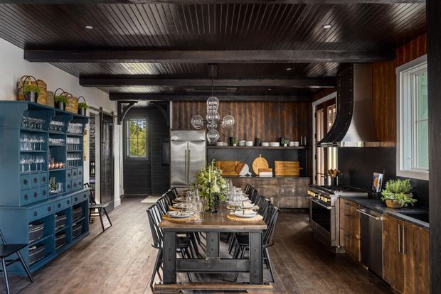 Dark, richly detailed kitchen and dining space with a long timber dining table set for entertaining. The room features dark timber ceiling boards and beams, timber flooring, black cabinetry and benchtops, a large sculptural rangehood, open shelving, a blue built-in display cabinet, stainless-steel refrigeration, and warm wood wall panelling.