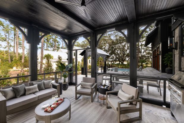 Covered outdoor entertaining and lounge area with a timber-framed, screened enclosure, grey upholstered sofa and armchairs, an oval coffee table, built-in barbecue, and views toward a paved terrace and swimming pool. The space has a refined contemporary resort-like character with dark structural framing and light grey finishes.