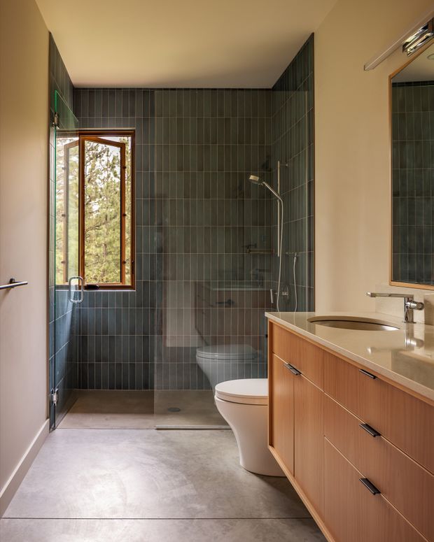 A contemporary bathroom with a walk-in glass shower, wall-hung toilet, freestanding-style toilet, single integrated basin, and extensive warm timber vanity cabinetry. The shower walls are clad in narrow vertical dark green-grey tiles, with a timber-framed opening window overlooking trees. A large mirrored cabinet and slim linear wall light are visible above the vanity. The room has a calm, refined palette of dark tile, warm wood, white sanitaryware, and pale grey flooring.