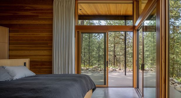 A warm, wood-lined bedroom with a partially visible bed in the foreground and expansive timber-framed glazing opening toward a wooded landscape. The room features horizontal timber wall boards, a timber ceiling, tall pleated curtains, and a polished grey concrete floor. The glazing includes a high clerestory-like transom and large sliding or folding doors, creating a strong indoor-outdoor connection.