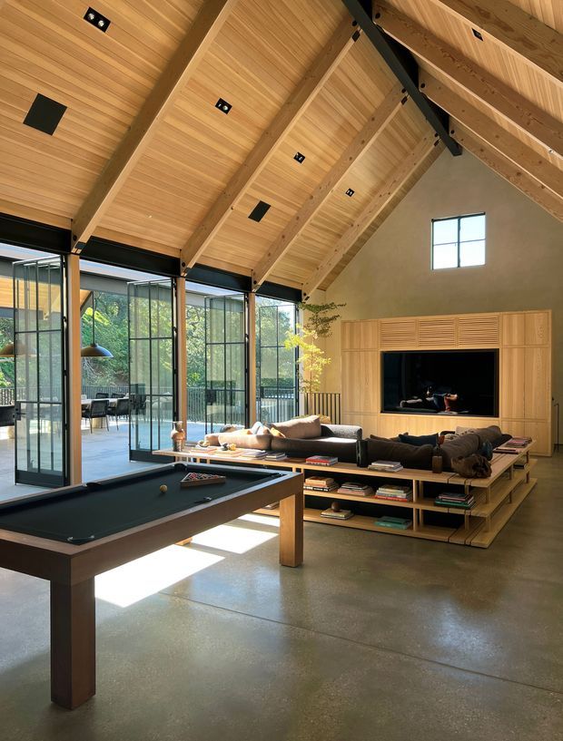 Spacious contemporary living and recreation area with a high pitched timber-lined ceiling, exposed timber and black steel beams, full-height industrial-style glazed doors, and polished concrete flooring. A pool table occupies the foreground, while a large sectional sofa, open timber shelving, and bespoke timber media cabinetry surround a fireplace or television recess. The glazing opens toward an outdoor terrace and leafy setting.