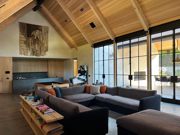 A spacious open-plan living room with a dramatic pitched, timber-lined ceiling and exposed timber rafters. A large dark sectional sofa surrounds a low timber coffee table, while a built-in timber kitchen and joinery sit at the rear. Floor-to-ceiling black-framed glazing opens the room visually to a bright courtyard or outdoor dining area. The room also features a large sculptural wall artwork and a warm, sophisticated material palette.