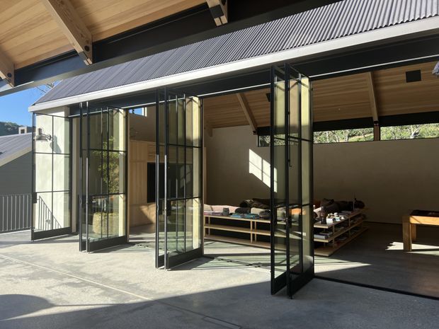 Open-sided contemporary pavilion or extension with extensive black-framed glazed folding doors, a high timber-lined ceiling, exposed dark structural beams, and a corrugated metal roof. The space opens onto a concrete terrace and contains built-in timber seating and shelving, with a calm indoor-outdoor arrangement and strong sunlight.