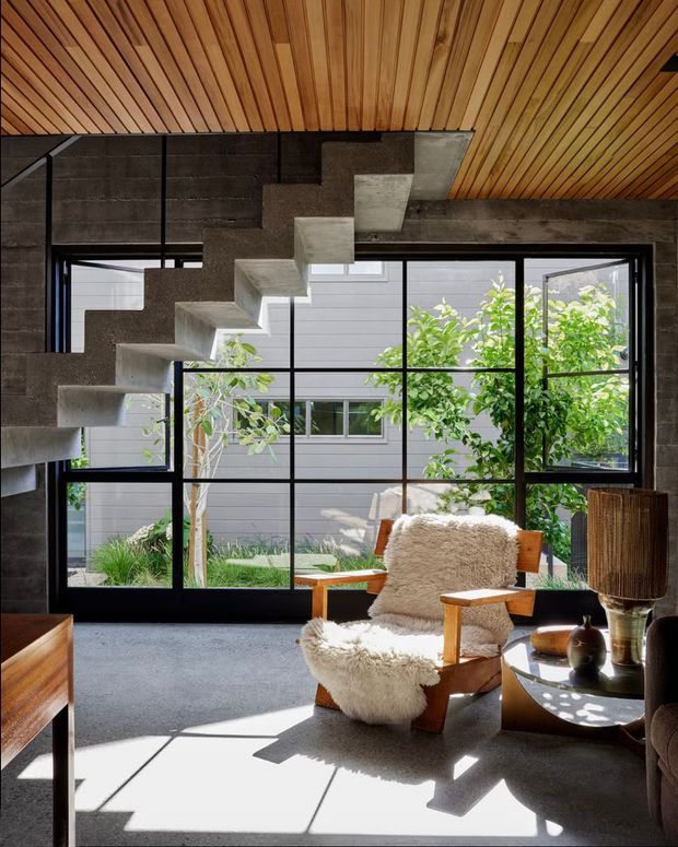 A sunlit modern living room featuring a dramatic exposed concrete stair with open risers, a warm linear timber-clad ceiling, large black-framed floor-to-ceiling glazing, and a sculptural timber lounge chair upholstered in cream sheepskin. The room opens visually to a planted courtyard and is furnished with a dark side table and contemporary lighting.