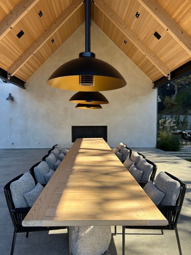 A long communal dining table with upholstered dining chairs sits beneath three oversized black pendant lights in a dramatic vaulted dining pavilion. The room features exposed timber roof framing and timber ceiling boards, smooth warm plaster walls, a dark built-in fireplace, polished concrete flooring, and open glazing to a landscaped outdoor area.