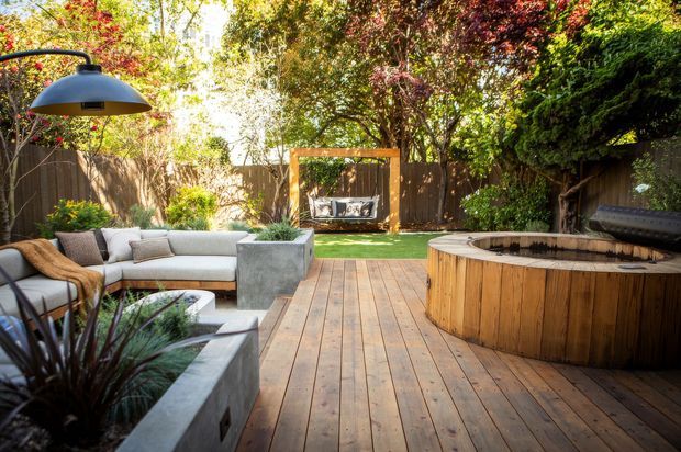 A sunny landscaped backyard featuring a timber deck, built-in outdoor lounge seating, a concrete planter, a large round timber-clad spa or hot tub, and a swing hanging from a simple timber frame. Mature planting and timber fencing create a private garden setting, with a sculptural outdoor lamp overhead.