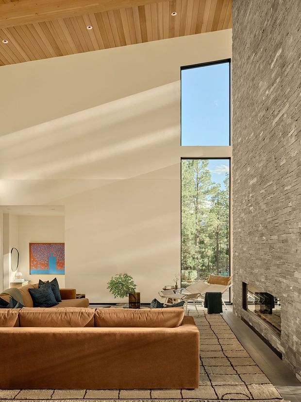 A spacious, warm-toned living room with a very high ceiling, a full-height textured stone fireplace wall, tall vertical glazing, and a timber-lined ceiling with recessed downlights. A large caramel leather sectional faces the fireplace, accompanied by a neutral patterned rug, lounge chairs, a small coffee table, and restrained decorative planting. Sunlight filters through the tall windows, creating a calm, refined atmosphere and connecting the room visually with the surrounding trees.